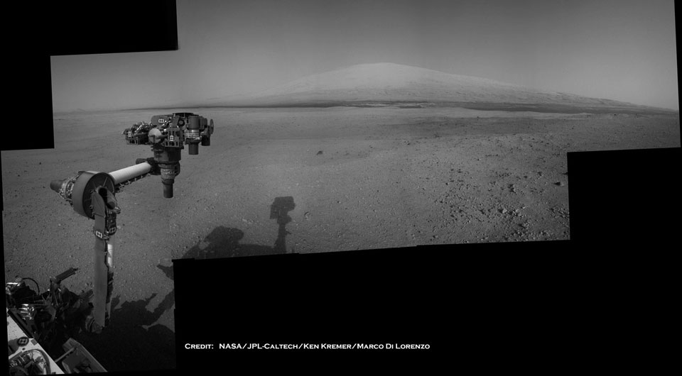 Ein Instrumentenarm des Marsrovers Curiosity ragt ins Bild, dahinter breitet sich eine steinige Ebene aus. Am Horizont ragt ein Berg in die Höhe.