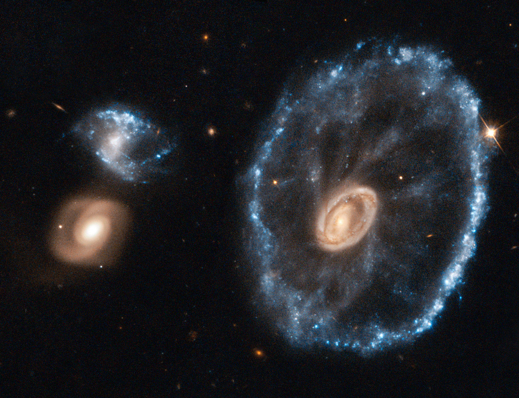 Die drei Galaxien im Bild wirken verzerrt. Rechts ist eine Galaxie, die im inneren Kern gelb leuchtet und sehr eng gewickelte Spiralarme hat. Außen herum verläuft ein blauer heller Kreis aus Sternen. Zwischen dem Kern und dem Ring verlaufen zarte Speichen. Links sind zwei viel kleinere Galaxien, die untere leuchtet gelblich und wirkt strukturiert, die obere ist verzerrt und leuchtet blau.