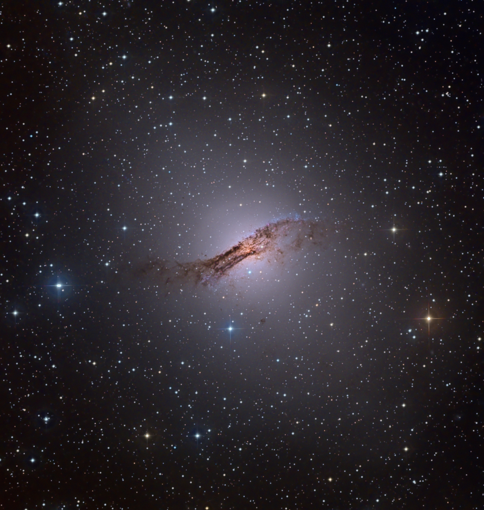 Centaurus A im Sternbild Zentaur ist die nächstgelegene aktive Galaxie in Erdnähe. Im Bild ist sie rund und leuchtet diffus. Davor verläuft ein breites, sehr markantes dunkles Staubband.