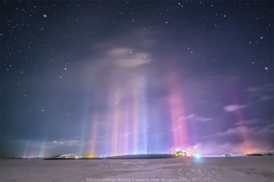 Viele dünne Lichtsäulen verbinden eine verschneite Landschaft mit einem sternenklaren Himmel. Der Große Wagen und weitere Sternbilder sind hinter den bunten Säulen zu sehen.