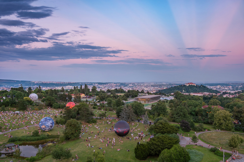 Das Bild zeigt einen Park mit vielen Menschen, in dem riesige Kugeln aufgestellt sind, sie repräsentieren Sonne, Erde, Mond und den Sternenhimmel. Über einem Schloss im Hintergrund auf einem Hügel laufen rosarote Gegendämmerungsstrahlen über der Festung Špilberk zusammen.