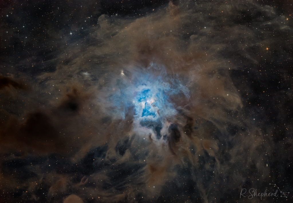 Mitten in einer bräunlichen Wolke leuchtet ein blauer Nebel. Staub streut blaues Licht effizienter als rotes Licht.