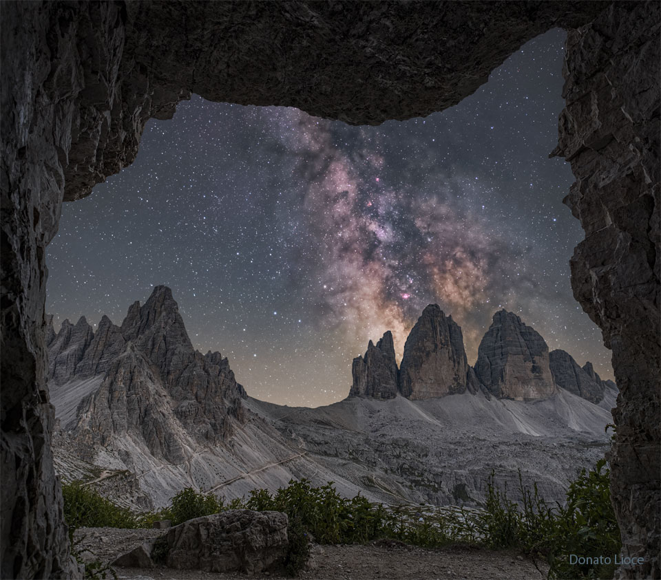 Von einem Überhang im Gebirge aus fällt der Blick auf eine Berglandschaft mit den berühmten Drei Zinnen in den Dolomiten. Über den Zinnen steigt die Milchstraße senkrecht auf.