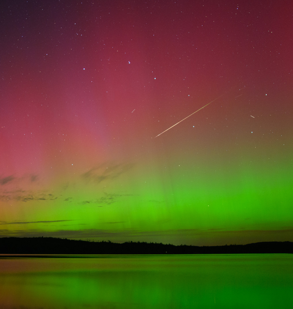 Über einem See schimmern unten grüne und oben rote Polarlichter. Oben am Himmel steht der Große Wagen, durch den Kasten zischt ein Meteor der Perseiden.