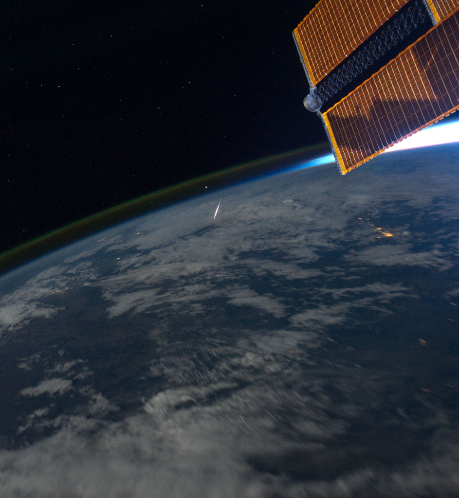 Rechts über der Erde ragt ein Solarpaneel der ISS ins Bild. Die Erde füllt die untere Bildhälfte, oben ist grünes Nachthimmellicht am Erdrand. In der Mitte leuchtet ein Splitter, es ist ein Meteor der Perseïden.