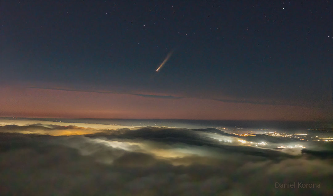 Aus einem Wolkenmeer ragen ein paar Hügel hervor, von unten leichten Lichter an die Wolkendecken, hinten sind Stadtlichter zu sehen. Über allem schwebt der Komet suchinshan–ATLAS mit einem prächtigen Schweif.