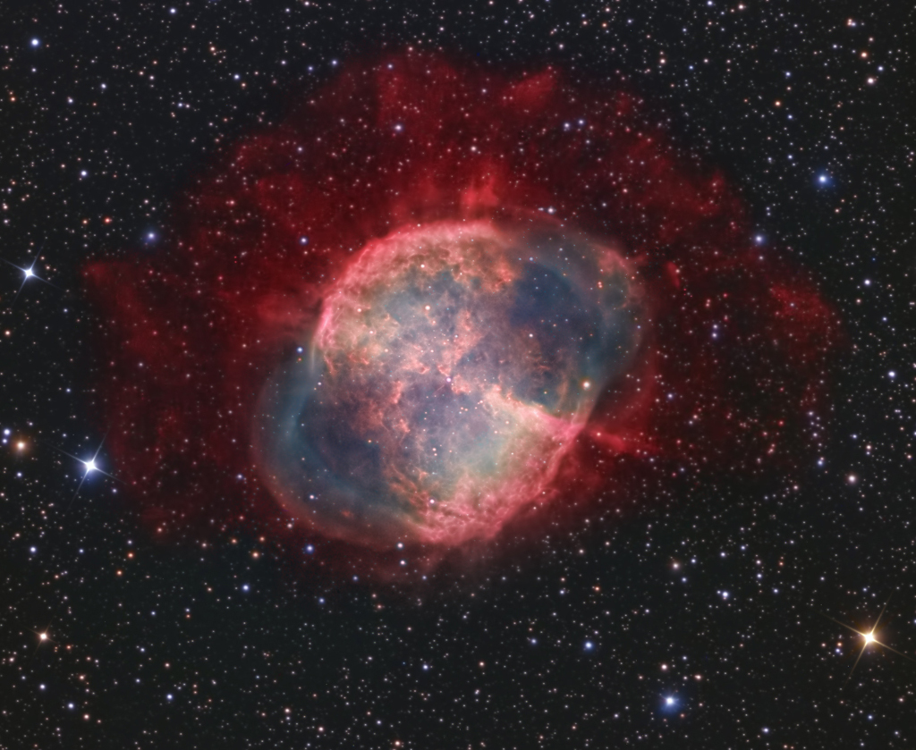 Der Hantelnebel im Sternbild Füchslein ist als M27 katalogisiert. Im Bild hat er rote Ränder und einige rot leuchtende Flecken vor zartblau schimmerndem Hintergrund.