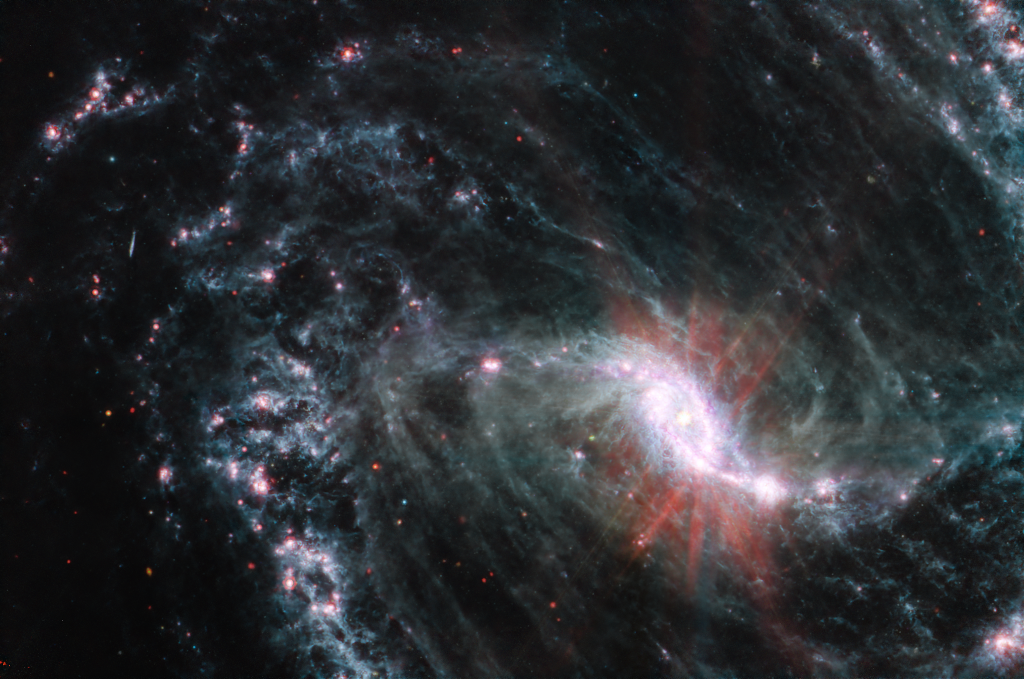 Vor dunklem Hintergrund sind in hellen nahezu weißen Farbtönen der zentrale Balken der Galaxie und die Spiralarme zu erkennen. Diese sind mit rötliche leuchtenden Flecken gesprenkelt. Im Zentrum der Galaxie leuchtet es hell rot. Ein Klick auf das Bild lädt die höchstaufgelöste verfügbare Version.