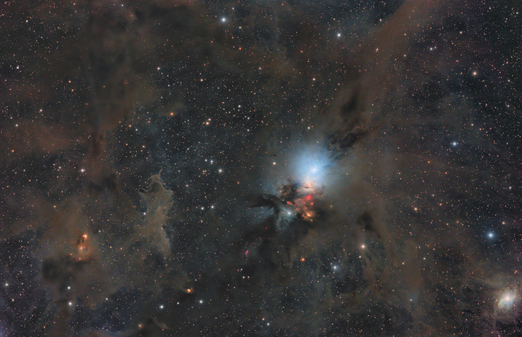 Mitten in braun-grauen Nebelwolken strahlt ein blauer Reflexionsnebel, darunter leuchten ein paar rot leuchtende Stellen aus dunklen Nebeln hervor. Zwischen den braunen Nebelfetzen leuchten dicht verteilte Sterne.