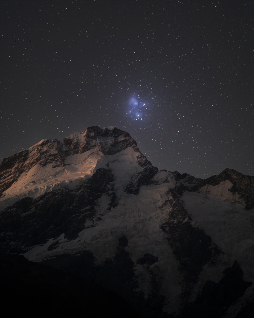 Über einem verschneiten Berg geht der Sternhaufen der Plejaden auf. M45 steht im Sternbild Stier. Sie sind von einem blauen Reflexionsnebel umgeben.