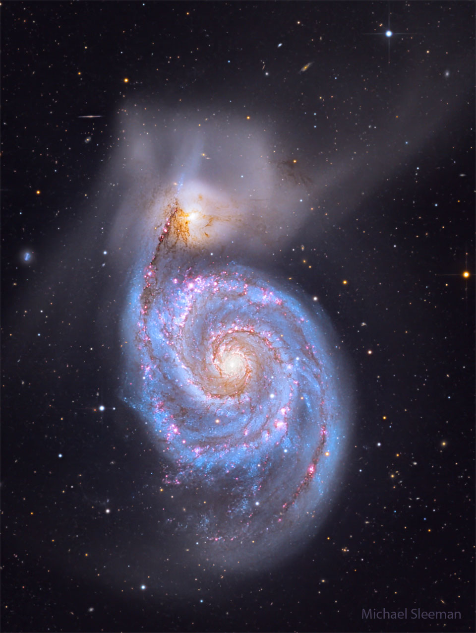 Über der markanten Spiralgalaxie M51, die hier blau und rot leuchtet und einen gelblichen Kern hat, liegt eine kleinere Begleitgalaxie, von der viele nebelige Strukturen ausgehen.