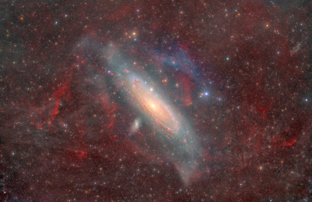 Die Andromeda-Galaxie wirkt hier anders als auf den meisten Bildern. Der Hintergrund ist mit rötlichen Schleiern und Nebeln bedeckt. Die Spiralarme sind von rötlichen Sternhaufen gesäumt.